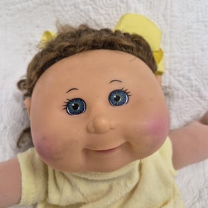 Peut inclure: Une poupée au visage rond couleur pêche, aux yeux bleus et aux cheveux en laine brune ornés d'un nœud jaune. La poupée a des joues roses et porte un haut jaune clair. Elle présente un nez arrondi et un léger sourire bouche fermée.