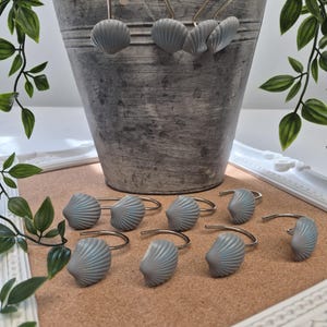 May include: A set of shower curtain hooks featuring grey, fan-shaped seashell ornaments attached to silver-toned metal rings. The shells have a ribbed texture and are arranged in a group, with some hanging from a metal bucket and others lying flat.