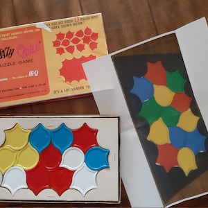 Może przedstawiać: W pudełku vintage KWAZY QUILE znajduje się gra logiczna z 13 kolorowymi elementami. Pudełko ma ż&oacute;łte tło z czerwonym i czarnym tekstem. Grę stworzyła firma Kenner, tw&oacute;rcy IQ. W pudełku znajdują się instrukcje i wyzwanie, aby dopasować elementy do obszaru pokazanego poniżej.