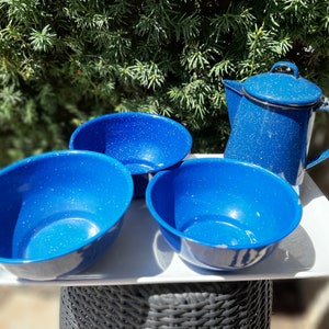 May include: A blue speckled enamelware camping set, comprising three bowls and a coffee pot with lid and handle. The items are arranged on a white tray, with a green leafy backdrop. The bowls and pot are blue.