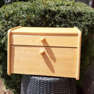 May include: A light wood bread box with two drawers and rounded edges. The bread box is sitting on a black wicker stool.