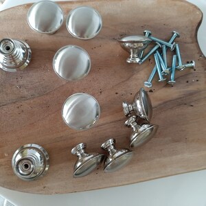 K&ouml;nnte beinhalten: Eine Sammlung von silbernen Schrank- und Schubladenkn&ouml;pfen mit Schrauben. Die Kn&ouml;pfe sind rund und haben eine glatte Oberfl&auml;che. Die Griffe sind oval und haben eine leicht strukturierte Oberfl&auml;che.