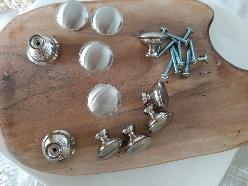 K&ouml;nnte beinhalten: Eine Sammlung von silbernen Schrank- und Schubladenkn&ouml;pfen mit Schrauben. Die Kn&ouml;pfe sind rund und haben eine glatte Oberfl&auml;che. Die Griffe sind oval und haben eine leicht strukturierte Oberfl&auml;che.