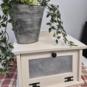 May include: A cream-coloured wooden bread box with a drop-down front door featuring a central glass panel etched with a floral design. The door has a black round knob and two black metal hinges. A grey metal bucket with a green plant sits on top.