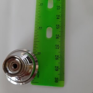 K&ouml;nnte beinhalten: Ein runder Schrankknopf in Silber mit einer erhabenen Mitte und einem kleinen Loch in der Mitte. Der Knopf wird an einem gr&uuml;nen Lineal gemessen.