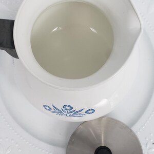 Op de afbeelding: Witte theepot van keramiek met een blauw bloemenmotief en een deksel van roestvrij staal. De theepot staat op een wit kantje.