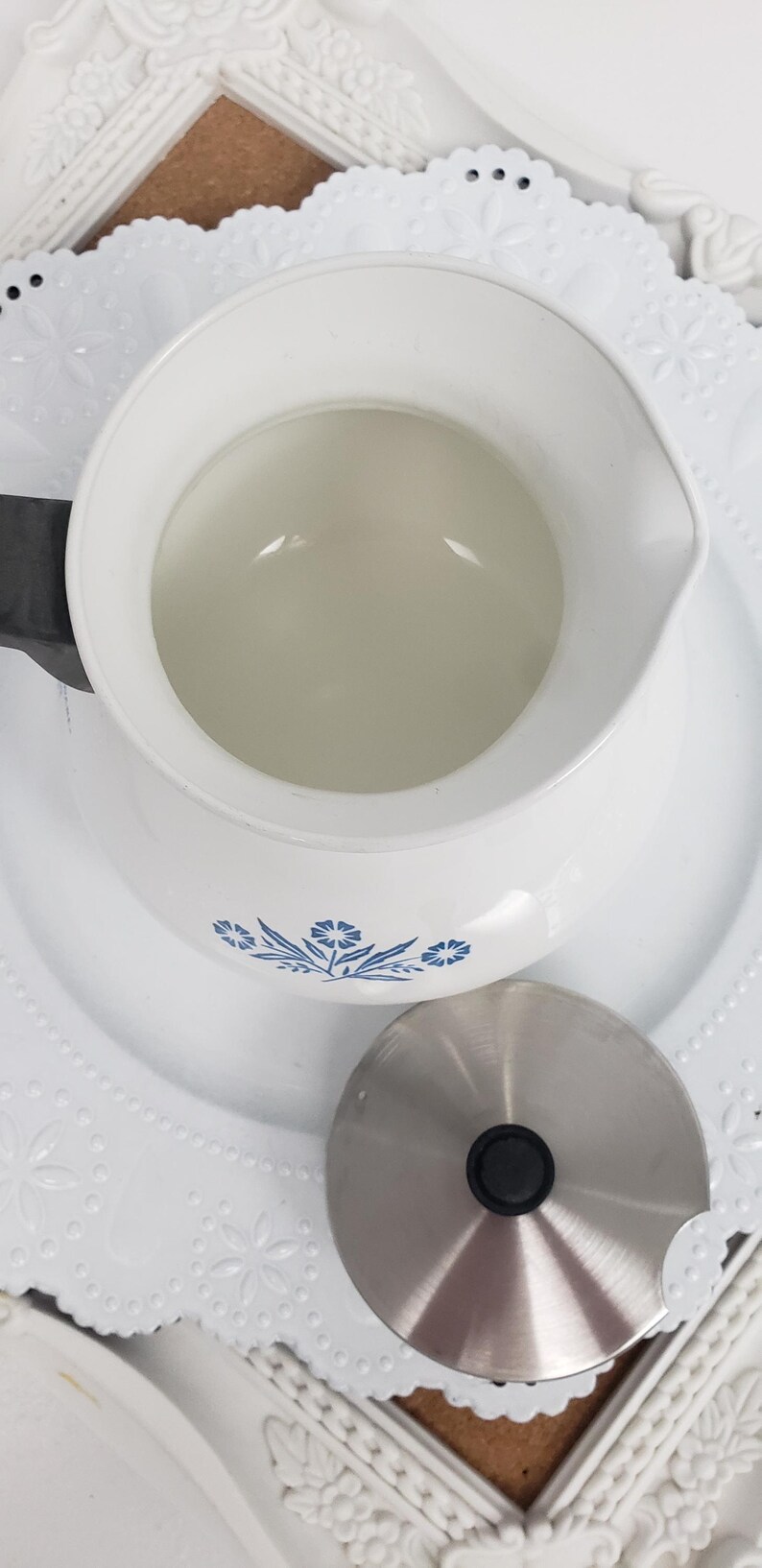 Op de afbeelding: Witte theepot van keramiek met een blauw bloemenmotief en een deksel van roestvrij staal. De theepot staat op een wit kantje.