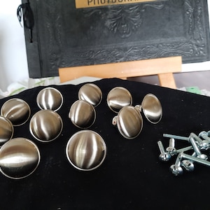 May include: A set of ten silver-toned round cabinet knobs with a shiny finish. The knobs are arranged in a semi-circle on a black surface. There are also ten silver screws included.