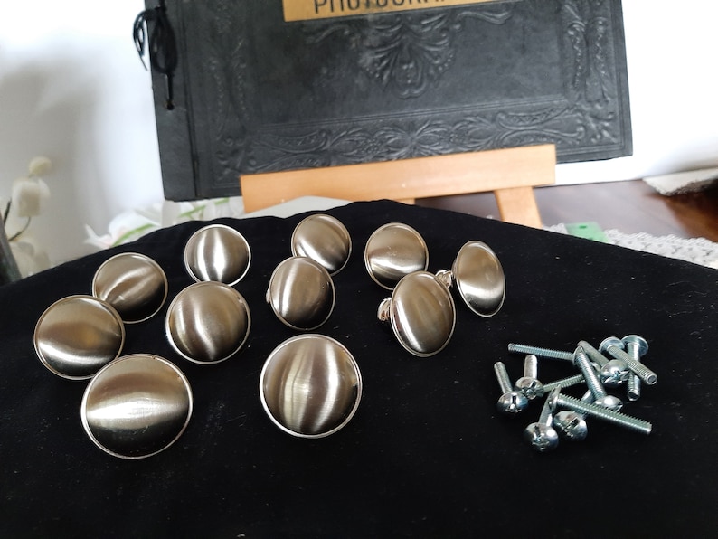 K&ouml;nnte beinhalten: Ein Satz mit zehn runden Schrankkn&ouml;pfen in Silber mit gl&auml;nzender Oberfl&auml;che. Die Kn&ouml;pfe sind in einem Halbkreis auf einer schwarzen Oberfl&auml;che angeordnet. Es sind auch zehn silberne Schrauben enthalten.