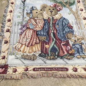 May include: A tapestry featuring two teddy bears dressed in red, blue, and plaid. The bears are standing on a wooden walkway with a white picket fence. The tapestry has a cream background with red hearts and the text "Just Between Us" and "Boyds Bears & Friends".