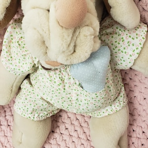 Puede incluir: Un peluche de perro con cuerpo color crema, orejas marrones y nariz rosa. Viste un vestido con estampado floral y un bolsillo azul. El juguete está sobre una manta rosa de punto grueso.