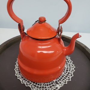 May include: A bright orange enamel teapot with a hinged lid and a single handle. The teapot is sitting on a white crocheted doily.