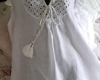 Peut inclure: Robe blanche en coton avec bordure en dentelle ajourée et un lien à glands au niveau du décolleté. La robe a des manches courtes bouffantes et une silhouette ample et fluide.