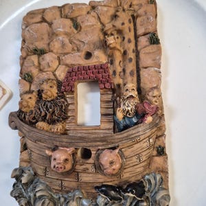 May include: A decorative light switch cover featuring a relief sculpture of Noah's Ark. The ark is filled with animals, including lions and pigs, and is set against a rocky backdrop. The cover is painted in earth tones with a detailed, textured finish.
