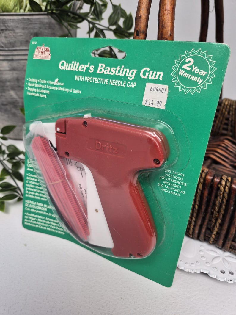 May include: A Dritz brand Quilter's Basting Gun with a protective needle cap, packaged on a green cardboard backing. The tool is dark red with a white trigger and handle. The packaging includes text, a 2-year warranty seal, and 500 tacks included.