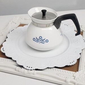 Op de afbeelding: Een witte koffiekan van keramiek met een blauw bloemenmotief en een zilveren deksel. De kan heeft een zwarte handgreep en staat op een wit kantonderzetter.