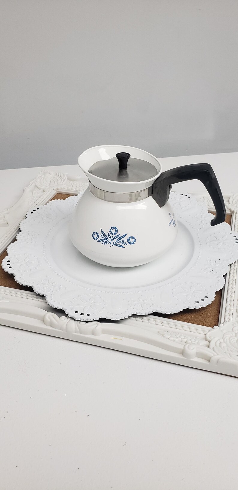 Op de afbeelding: Een witte koffiekan van keramiek met een blauw bloemenmotief en een zilveren deksel. De kan heeft een zwarte handgreep en staat op een wit kantonderzetter.