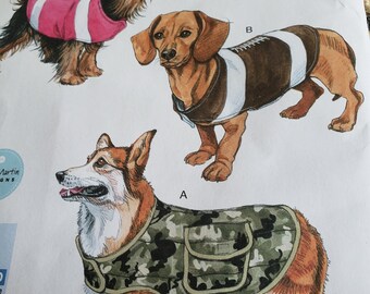 May include: A sewing pattern for dog clothes, featuring three different dog breeds wearing different outfits. The pattern includes a camouflage vest, a football jersey, and a pink jersey. The pattern is labeled "Style L9698, Size S-L, Bin #96".