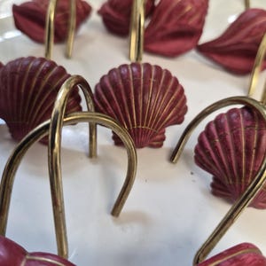 May include: A collection of shower curtain hooks featuring deep red, scalloped seashell designs with gold-toned accents. Each hook is attached to a curved, polished brass-coloured metal loop, designed for sliding onto a standard shower curtain rod.