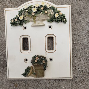 Könnte beinhalten: Ein weißer Lichtschalter aus Keramik mit einem Blumendesign. Die Abdeckung verfügt über ein erhabenes Design eines Blumentopfs mit Blumen und einer Vase mit Blumen.