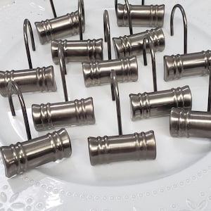 May include: A set of 12 silver shower curtain hooks with a decorative design. The hooks are arranged on a white plate with a lace pattern.