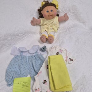 May include: A Cabbage Patch Kids doll with brown hair and a yellow bow, wearing a yellow top and white trousers. Included are a blue and white romper, a white onesie with monkey prints, a yellow blanket, a grey dummy, and a yellow note.