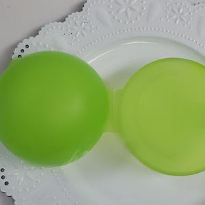 Op de afbeelding: Een limoengroene, ronde container met een bijpassend deksel, verbonden door een kleine brug. De container staat op een witte, decoratieve plaat met een geschulpte rand en een bloemmotief.