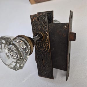 Op de afbeelding: Een deurbeslagset met een heldere, gefacetteerde glazen knop bevestigd aan een donkere, sierlijke metalen achterplaat met ingewikkelde bloemmotieven en een sleutelgat, verbonden met een rechthoekig ijzeren insteekslot.