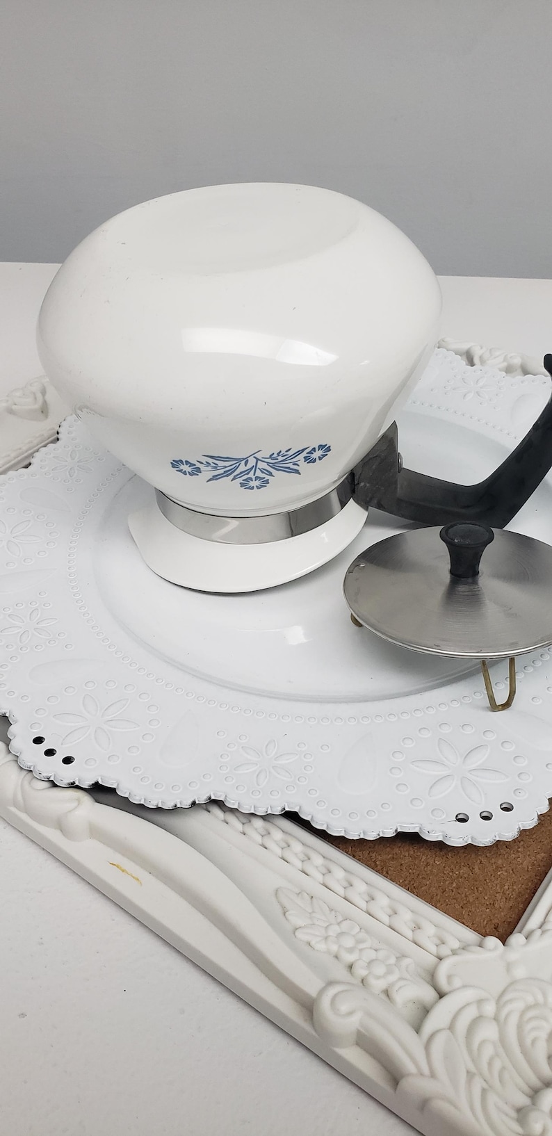 Op de afbeelding: Witte theepot met een blauw bloemenmotief en een zwarte handgreep. De theepot staat op een wit kantje.