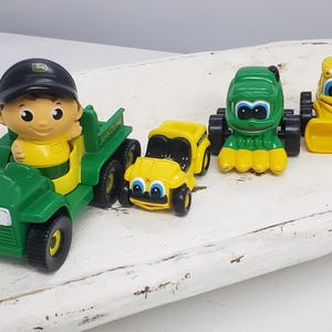 May include: A set of four green and yellow toy tractors. One tractor has a figure of a person in a green shirt and black hat driving it. The tractors have smiling faces and wheels.