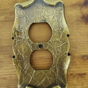 Op de afbeelding: Een gouden stopcontact afdekplaat met een decoratief, gestructureerd ontwerp. De afdekplaat heeft een licht sierlijke, gegolfde rand.
