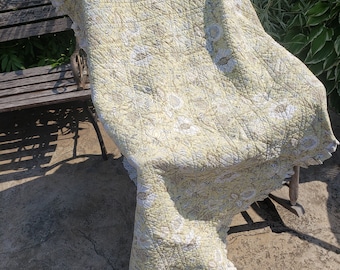May include: A yellow and white floral patterned quilt with a ruffled edge is draped over a wooden bench.