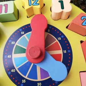 May include: A colourful wooden clock with a spinning hand and a dial marked with numbers from 1 to 12 and numbers from 5 to 55. The clock has a yellow background and a blue and red hand.