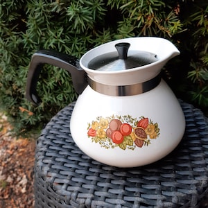 May include: White vintage teapot with a black handle and lid. The teapot features a colorful fruit and floral design. The teapot is sitting on a black wicker surface, with green foliage in the background.