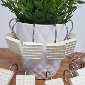 May include: Set of white shower curtain hooks with a square grid pattern. The hooks are hanging on a white ceramic planter with a green plant.