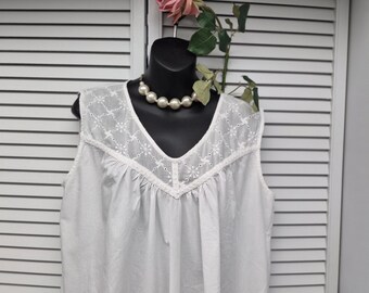 Peut inclure: Chemise de nuit blanche sans manches avec des détails brodés autour de l'encolure. La chemise de nuit est présentée sur une tête de mannequin noire, ornée d'un collier de perles et d'une rose rose. L'arrière-plan est une porte à volets blancs.