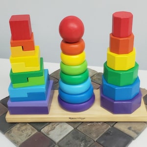 May include: Wooden stacking toys in a rainbow of colors. The set includes three different designs: a square block tower, a round ring tower, and a hexagonal block tower. The toys sit on a wooden base.