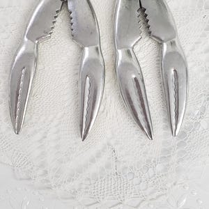 May include: Two silver crab crackers with a claw-like design, featuring serrated edges for gripping. The tools are displayed on a white, decorative plate with a floral pattern.