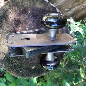 Pode incluir: Um conjunto de maçanetas de porta vintage com maçanetas de cerâmica preta e uma placa de metal com um buraco para chave. As maçanetas são presas a uma haste de metal e a placa é presa a uma superfície de madeira.