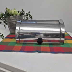 May include: A polished stainless steel bread bin with a curved, roll-top lid and a small, round black knob. The container features a reflective metallic surface and a rectangular base, providing a sleek and functional storage solution for kitchen worktops.