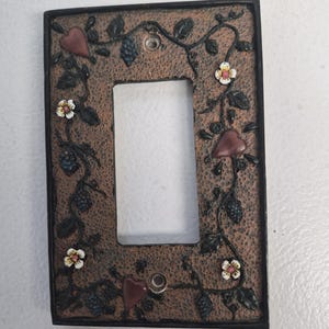 May include: Decorative light switch cover with a textured brown background and a black border. The cover is adorned with vines, flowers, and heart-shaped accents. The flowers have white petals and yellow centers. The design includes clusters of blue grapes.