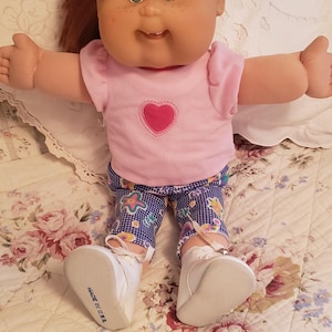 Op de afbeelding: Een Cabbage Patch Kids pop met bruin haar en groene ogen, die een roze shirt met een hartje erop draagt, een blauw-wit geblokte broek en witte schoenen. De pop zit op een bedsprei met bloemenprint.