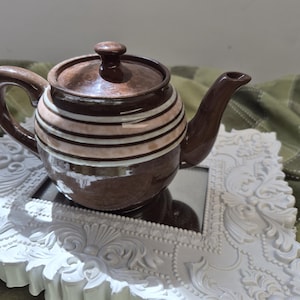 May include: A brown ceramic teapot with a glossy finish, featuring a lid with a small knob, a curved handle, and a spout. The body of the teapot is decorated with horizontal white and light pink stripes around the centre, creating a distinct contrast.