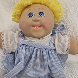 May include: A doll with bright yellow yarn hair, blue eyes, and two small front teeth. The doll wears a white short-sleeved shirt with lace trim on the sleeves and a light blue and white gingham pinafore dress with shoulder straps.