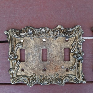 Könnte beinhalten: Eine goldfarbene Metall-Lichtschalterplatte mit einem verzierten Design mit Rosen und Schnörkeln. Die Platte hat drei Kippschalter.