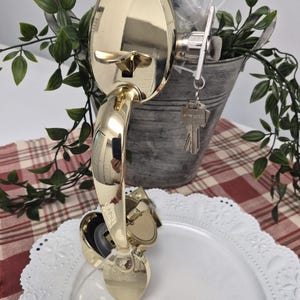 May include: A polished gold-tone metal door handle set featuring an arched grip, a thumb-press latch, and a circular backplate. A silver-tone metal key is inserted into the lock cylinder on the side, and the hardware is connected by a clear plastic tie.