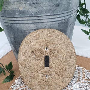 Puede incluir: Una cubierta de interruptor de luz redonda de cerámica de color beige con un diseño de cara de sol. La cubierta tiene una abertura rectangular para el interruptor y dos pequeños agujeros. Un cubo de metal galvanizado con vegetación está en el fondo.