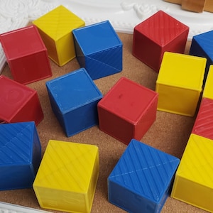 May include: A collection of colorful toy blocks in red, yellow, and blue. The blocks are arranged on a corkboard within a white decorative frame. Some blocks have diagonal line patterns.