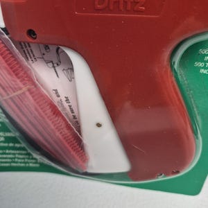 May include: A red and white Dritz tagging gun encased in clear plastic packaging. The tool features a prominent red handle and a white trigger mechanism. The packaging includes visible text and instructions for use, with a red plastic fastener strip.