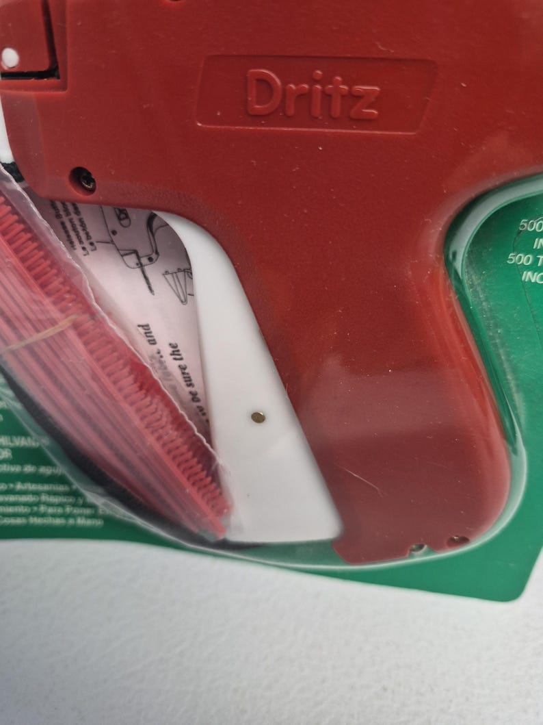 May include: A red and white Dritz tagging gun encased in clear plastic packaging. The tool features a prominent red handle and a white trigger mechanism. The packaging includes visible text and instructions for use, with a red plastic fastener strip.